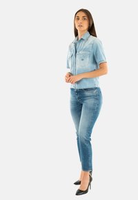Camisa de mezclilla ligera de manga corta con dos bolsillos en el pecho, combinada con jeans ajustados de tono azul medio y zapatos de tacón negro.