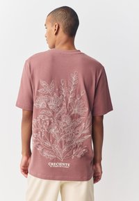 Pink cotton t-shirt featuring white floral line art and "CRECIENTE EL CAMPO DE FLORES" text at the bottom, short sleeves, relaxed fit.