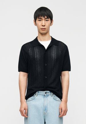 Chemise noire à manches courtes avec des panneaux à motif texturé, col et boutonnage à l'avant. Associée à un short en denim bleu clair. Fond uni.
