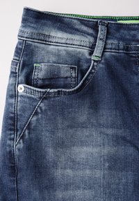 Tela de denim azul oscuro con un aspecto descolorido, que presenta un pequeño bolsillo frontal, una costura de acento verde y herrajes metálicos plateados.