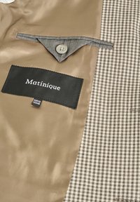 Brun satinfoder med en grå tygficka, med en svart etikett med vit text som säger "Matinique" och ett hundtandsmönster på höger sida.