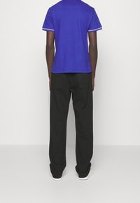 Modrá košile s krátkým rukávem a bílými pruhovanými akcenty, spárovaná s černými nohavicemi. Oblečení má uvolněný střih a minimální detaily.