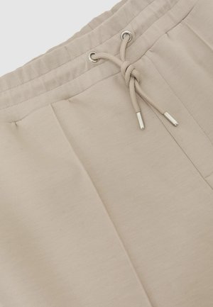 Pantalon de survêtement beige avec une taille élastique, une fermeture à cordon de serrage et une texture de tissu lisse. Comprend deux poches latérales et des coutures discrètes.