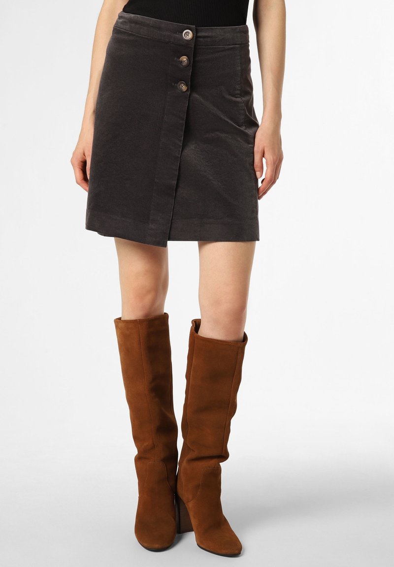 Femme portant une jupe noire boutonnée et des bottes en daim marron à talons montant jusqu'aux genoux, debout sur un fond blanc uni.