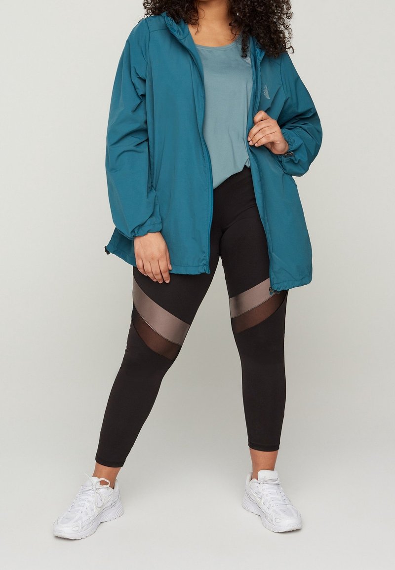 Teal let jakke over en lys blå top, parret med sorte leggings med gennemsigtige paneler. Hvide sneakers fuldender outfittet.
