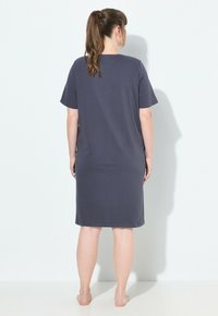 Donna con una coda di cavallo castana, sta scalza, di fronte a un muro bianco, indossando un vestito scuro grigio a maniche corte, largo e fino al ginocchio.