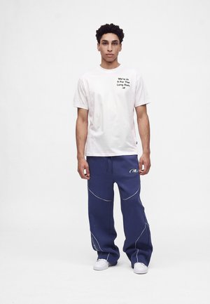 Νέος άνδρας στέκεται φορώντας λευκό T-shirt με κείμενο και μπλε φαρδιά αθλητική φόρμα με λευκά αθλητικά παπούτσια σε μονόχρωμο φόντο.
