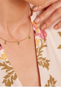 Collar de cadena de oro con tres pequeñas gemstones verdes, llevado en el cuello, mano con esmalte de uñas nude tocando una prenda blanca con patrón floral.