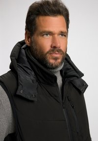Schwarze Steppweste mit hohem Kragen und Druckknöpfen, sowie Reißverschlusstaschen, getragen über einem grauen Pullover. Sie besteht aus glattem, isoliertem Stoff.