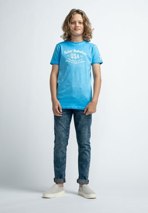 Camiseta de algodón azul con texto blanco, diseño desgastado, mangas cortas. Combinada con jeans de mezclilla oscuros, enrollados, y zapatos slip-on gris claro.