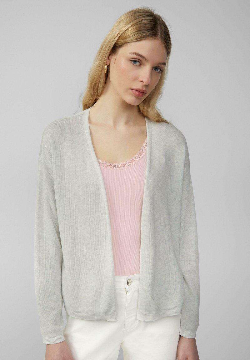 Cardigan gris clair avec ouverture devant, texture douce, manches longues et poignets côtelés sur un débardeur rose clair. Design simple et décontracté.