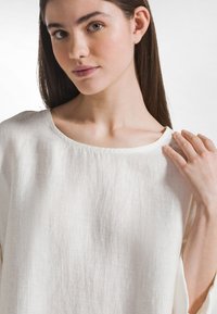 Hvid linnedtop med rund halsudskæring, løstsiddende pasform og korte ærmer. Stoffet har en subtil tekstur og let glans.