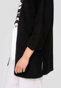 Schwarze Strickjacke mit lockerer Passform, drei Viertelärmeln, seitlichen Schlitzen und strukturierter Oberfläche. Über einem weißen Kleidungsstück getragen.