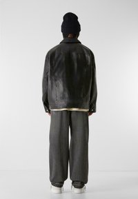 Schwarze Lederjacke mit lockerer Passform, sichtbaren Nähten und zwei geknöpften Manschetten; kombiniert mit lockeren grauen Hosen und weißen Sneakers.