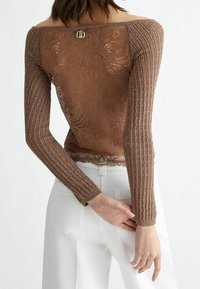 Ruskea pitkähihainen toppi, jossa on neulotut hihat ja kukkakuvioinen pitsi takana, yhdistettynä korkeavyötäröisiin valkoisiin housuihin, nähtynä takaa.