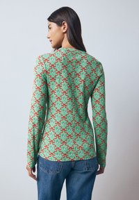 Haut vert à manches longues avec un motif répétitif rouge et blanc, fermeture éclair au dos et texture côtelée. Associé à un jean bleu.