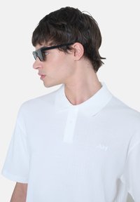 Polo shirt bianca a righe con colletto, dotata di una patta con tre bottoni e un piccolo logo ricamato. Indossata con occhiali da sole neri a forma rettangolare.