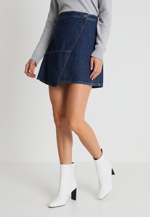 Femme portant une jupe portefeuille en denim bleu foncé, un pull gris clair à manches longues, et des bottines blanches à talons hauts, marchant devant un fond uni.