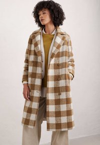Braun-weiß karierter Wollmischungsmantel mit einem eingeschnittenen Kragen, doppelreihigen Knöpfen und Seitentaschen, über einem senfgelben Pullover getragen.