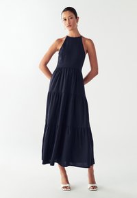 Žena stojaca s rukami za chrbtom, oblečená v bezrukávových námorníckych tiered maxi šatách a bielych sandáloch s otvorenou špičkou na bielom pozadí.