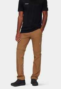 Muškarac u crnoj Mammut majici i smešnim konvertibilnim hlačama stoji s jednom rukom u džepu i nosi crne cipele.