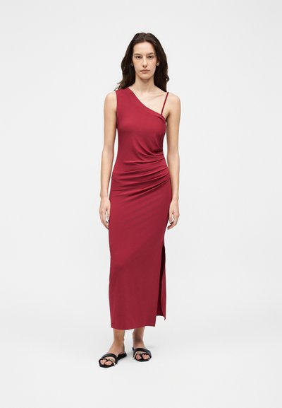 Femme portant une robe midi asymétrique sans manches rouge foncé avec une fente latérale, debout devant un fond clair uni.