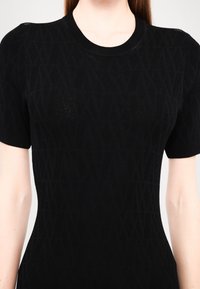 Femme portant un pull noir ajusté à manches courtes avec un motif texturé subtil et un col rond, montrant le haut du torse et les bras.