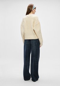 Suéter grueso de punto de cable con escote en V y corte oversized, combinado con jeans oscuros de pierna ancha con bolsillos traseros visibles.