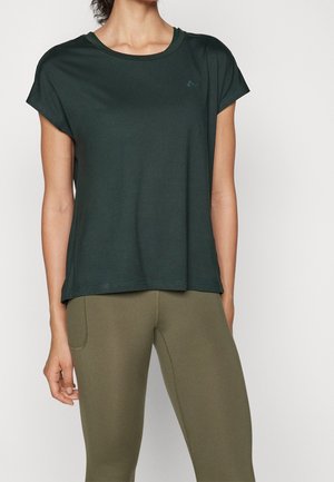 Vrouw draagt een donker groen T-shirt met korte mouwen en olijfgroene aansluitende broek, staand tegen een effen witte achtergrond.