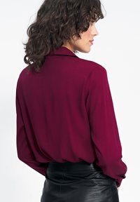 Bordowa bluzka z długimi rękawami i wciętym kołnierzykiem, wykonana z gładkiego, lekkiego materiału. Noszona z elegancką, czarną skórzaną spódnicą.