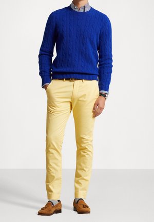 Hombre con un suéter azul de punto trenzado, camisa a rayas, pantalones amarillos, un reloj y mocasines marrones con borlas, de pie con una mano en el bolsillo.