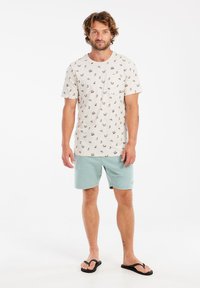 Camiseta beige claro con pequeños patrones de anclas, combinada con pantalones cortos verde menta. El modelo lleva chanclas negras; estilo casual y relajado.