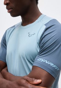 Άνδρας που φοράει αθλητική μπλούζα κοντομάνικη σε ανοιχτό μπλε και γκρι χρώμα με λογότυπο πάνθηρα στο στήθος και το κείμενο "DYNAFIT" στο μανίκι, με σταυρωμένα τα χέρια.