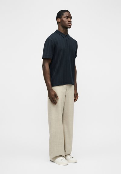 Άνδρας που στέκεται φορώντας σκούρο υφασμάτινο πόλο με κοντά μανίκια, μπεζ παντελόνι φαρδιάς γραμμής και λευκά αθλητικά παπούτσια, μπροστά σε ένα απλό φωτεινό φόντο.