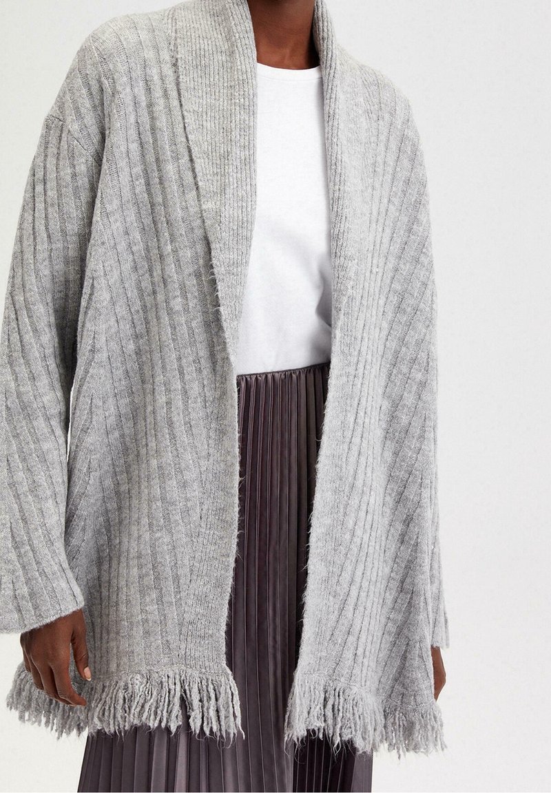 Šedý žebrovaný kardigan s třásněmi na okrajích, vyrobený z texturované pleteniny. Nasaďte ho přes bílou košili a plisovanou sukni pro ležérní vzhled.
