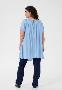 Blouse bleu clair à manches courtes retroussées et détail en forme de trou de serrure au dos, portée avec un jean évasé bleu foncé. Tissu doux et respirant.