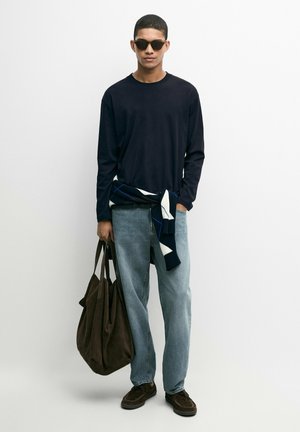 Junger Mann trägt schwarzes Langarmshirt, hellblaue Jeans, Sonnenbrille, dunkle Schuhe, hält große braune Tasche, Pullover um die Taille gebunden.