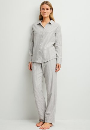 Vrouw staat blootsvoets, draagt lichtgrijs gestreept, lang mouw, met knopen gesloten pyjamashirt en bijpassende losse broek tegen een effen achtergrond.