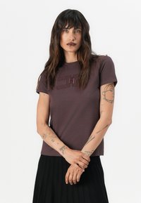 T-shirt en coton bordeaux à manches courtes avec un logo géométrique texturé sur le devant. Le modèle porte une jupe plissée noire.