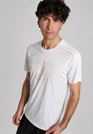 Uomo con capelli ricci che indossa una maglietta bianca con cuciture gialle e la scritta "LEONE 1947" sul petto, in piedi davanti a uno sfondo grigio.