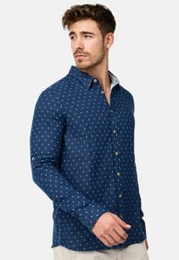 Camisa de botones azul marino con un sutil patrón de cuadrados, mangas largas y un cuello. Presenta botones marrones y una tela texturizada.