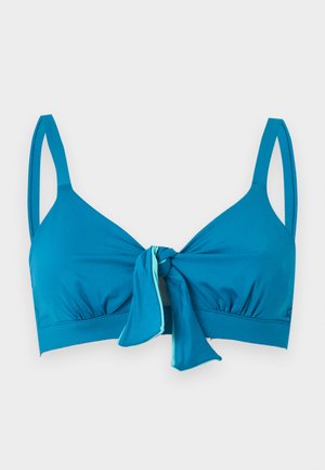 Jeune femme debout portant un bikini turquoise avec un haut noué devant et un bas noué sur les côtés, associé à des sandales noires à enfiler, sur un fond blanc uni.