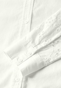 Camisa blanca de botones con una parte frontal lisa y un sutil patrón bordado en el puño enrollado. Tela de algodón suave y collar estándar.