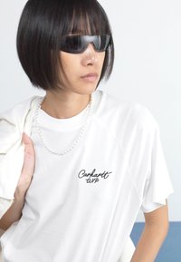 White short-sleeve t-shirt with black "Carhartt WIP" logo, paired with a pearl necklace and dark sunglasses, against a pale background.