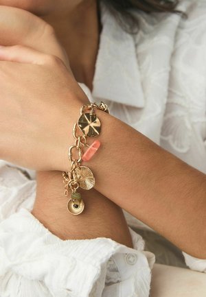 Guldmetal armbånd med forskellige charms, herunder flade skiver, en pink perle og en grøn sten. Kædelinks tilføjer struktur og dimension.