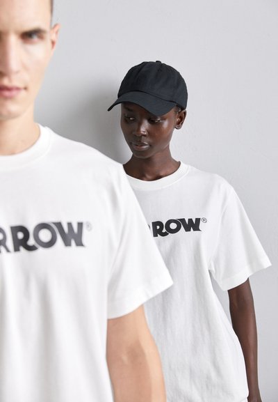 Białe bawełniane koszulki z dużym czarnym logo "ARROW" na przodzie, z czapką w kolorze czarnym noszoną przez modela w tle.