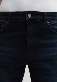 Granatowe dżinsy o gładkiej fakturze, z zapięciem na guzik z przodu i dwiema kieszeniami. Widoczne szwy wzdłuż szwów.