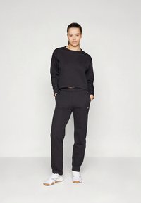 Svart cropped sweatshirt og svarte joggebukser laget av mykt stoff, med snøring i midjen og liten logo på siden. Brukt med hvite sneakers.