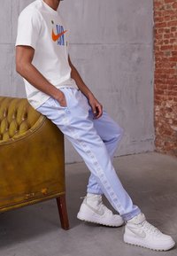 Bílé tričko Nike s barevným logem; světle modré tepláky se zipy po stranách; bílé vysoké tenisky; hnědé čalouněné kožené křeslo.