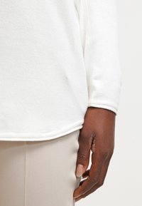 Maglione bianco a maniche lunghe con texture visibile e polsini a costine, abbinato a pantaloni beige su misura, mostrando un primo piano di una mano appoggiata sul tessuto.